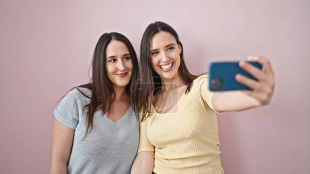 Foto de Dos mujeres sonriendo confiadas hacen selfie por teléfono inteligente sobre fondo rosa aislado - Imagen libre de derechos