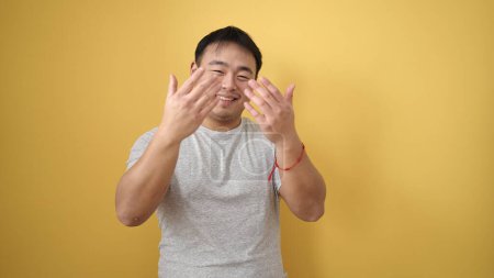 Téléchargez les photos : Jeune homme chinois souriant confiant faire geste à venir avec les mains sur fond jaune isolé - en image libre de droit