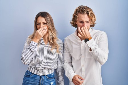 Téléchargez les photos : Jeune couple debout sur fond bleu sentant quelque chose de puant et dégoûtant, odeur intolérable, retenant souffle avec les doigts sur le nez. mauvaise odeur - en image libre de droit