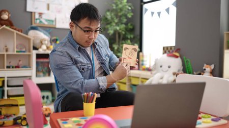 Téléchargez les photos : Jeune homme chinois enseignant préscolaire ayant une leçon de vocabulaire en ligne à la maternelle - en image libre de droit
