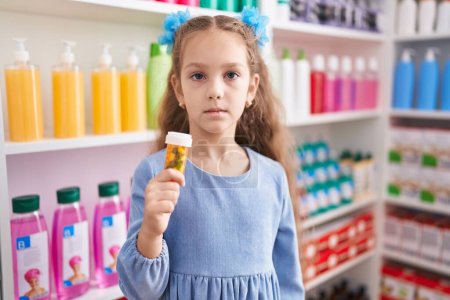 Photo for Young little girl holding pills at the pharmacy thinking attitude and sober expression looking self confident - Royalty Free Image