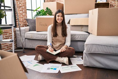 Téléchargez les photos : Jeune femme brune déménageant dans une nouvelle maison faisant des finances regardant positif et heureux debout et souriant avec un sourire confiant montrant les dents - en image libre de droit