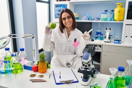 Téléchargez les photos : Jeune femme hispanique travaillant au laboratoire scientifique tenant des fruits souriant heureux pointant avec la main et le doigt vers le côté - en image libre de droit