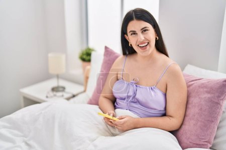 Photo for Young hispanic woman using smartphone sitting on bed at bedroom - Royalty Free Image
