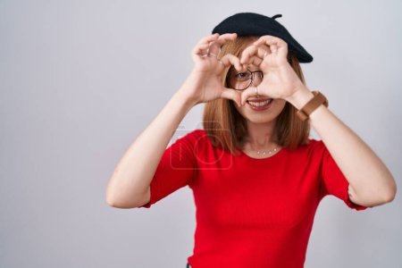 Photo for Young redhead woman standing wearing glasses and beret doing heart shape with hand and fingers smiling looking through sign - Royalty Free Image