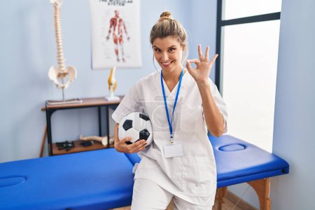 Téléchargez les photos : Jeune femme travaillant à la clinique de thérapie de football faire ok signe avec les doigts, sourire geste amical excellent symbole - en image libre de droit