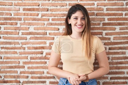 Téléchargez les photos : Jeune femme brune debout sur un mur de briques avec les mains jointes et les doigts croisés souriant détendu et joyeux. succès et optimisme - en image libre de droit
