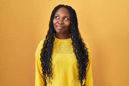 Foto de Mujer africana de pie sobre fondo amarillo sonriendo mirando hacia un lado y mirando hacia otro pensando. - Imagen libre de derechos