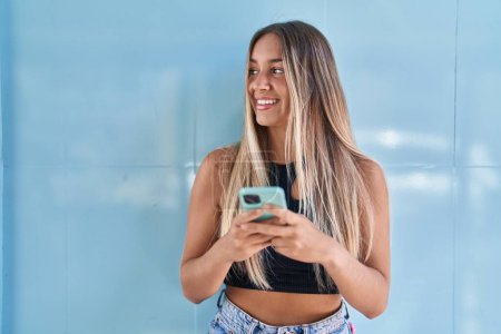 Téléchargez les photos : Jeune belle femme hispanique souriant confiant en utilisant smartphone sur fond bleu isolé - en image libre de droit