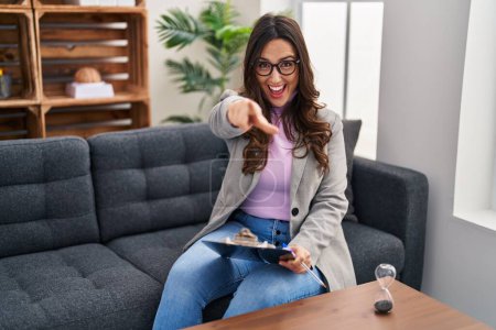 Téléchargez les photos : Jeune femme brune travaillant au bureau de consultation vous pointant du doigt et la caméra, souriant positif et joyeux - en image libre de droit
