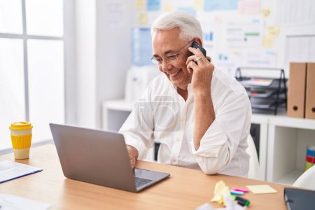 Téléchargez les photos : Homme d'affaires aux cheveux gris du Moyen-âge utilisant un ordinateur portable parlant sur smartphone au bureau - en image libre de droit