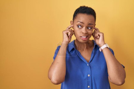 Photo for African american woman standing over yellow background covering ears with fingers with annoyed expression for the noise of loud music. deaf concept. - Royalty Free Image