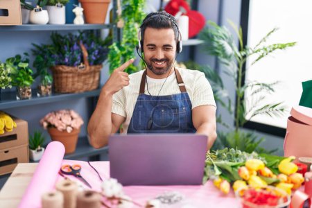 Téléchargez les photos : Homme hispanique avec barbe travaillant à la boutique de fleuristes en utilisant un ordinateur portable souriant heureux pointant avec la main et le doigt - en image libre de droit