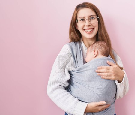 Téléchargez les photos : Jeune femme caucasienne tenant et portant bébé sur une fronde regardant positif et heureux debout et souriant avec un sourire confiant montrant des dents - en image libre de droit