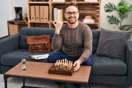 Photo for Middle age caucasian man playing chess sitting on the sofa pointing thumb up to the side smiling happy with open mouth - Royalty Free Image