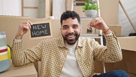 Téléchargez les photos : Jeune homme hispanique souriant confiant tenant tableau noir et clés à la nouvelle maison - en image libre de droit