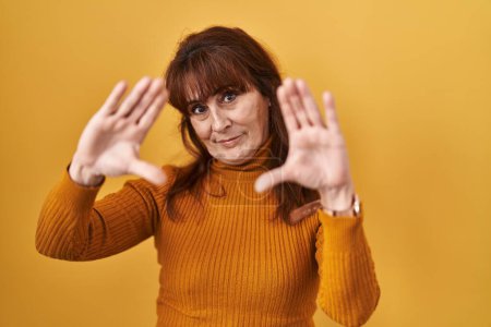Photo for Middle age hispanic woman standing over yellow background doing frame using hands palms and fingers, camera perspective - Royalty Free Image