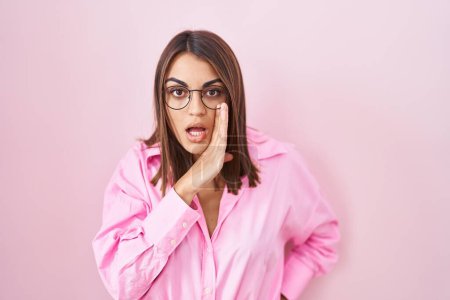Téléchargez les photos : Jeune femme hispanique portant des lunettes debout sur fond rose main sur bouche racontant rumeur secrète, chuchotant conversation malicieuse - en image libre de droit