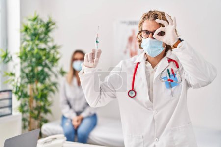 Photo for Blond man wearing doctor uniform and medical mask holding syringe smiling happy doing ok sign with hand on eye looking through fingers - Royalty Free Image
