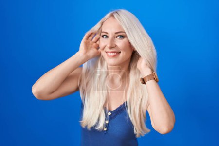 Téléchargez les photos : Femme blanche debout sur fond bleu essayant d'entendre les deux mains sur le geste de l'oreille, curieuse de commérages. problème auditif, sourd - en image libre de droit