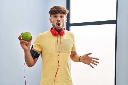 Photo for Arab man with beard wearing sportswear eating green apple scared and amazed with open mouth for surprise, disbelief face - Royalty Free Image