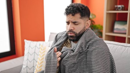 Foto de Joven hombre hispano sentado en el sofá cubriendo con manta para el frío en h - Imagen libre de derechos