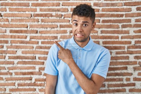 Photo for Brazilian young man standing over brick wall pointing aside worried and nervous with forefinger, concerned and surprised expression - Royalty Free Image