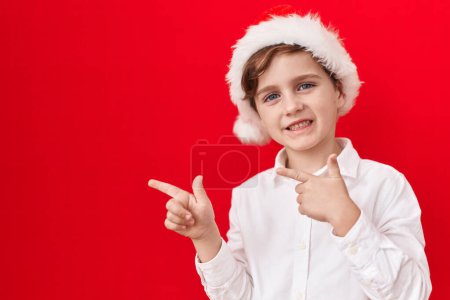 Photo for Little caucasian boy wearing christmas hat over red background smiling and looking at the camera pointing with two hands and fingers to the side. - Royalty Free Image