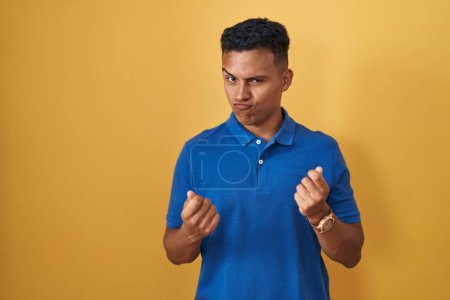 Foto de Joven hispano parado sobre fondo amarillo haciendo gesto de dinero con las manos, pidiendo pago de sueldo, negocio millonario - Imagen libre de derechos