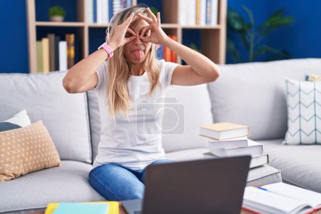 Photo for Young blonde woman studying using computer laptop at home doing ok gesture like binoculars sticking tongue out, eyes looking through fingers. crazy expression. - Royalty Free Image