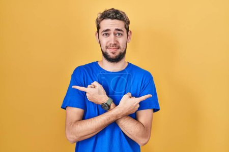 Photo for Hispanic man with beard standing over yellow background pointing to both sides with fingers, different direction disagree - Royalty Free Image