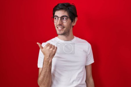 Foto de Joven hombre hispano de pie sobre fondo rojo sonriendo con la cara feliz mirando y señalando a un lado con el pulgar hacia arriba. - Imagen libre de derechos