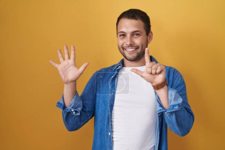 Foto de Hispanic man standing over yellow background showing and pointing up with fingers number seven while smiling confident and happy. - Imagen libre de derechos