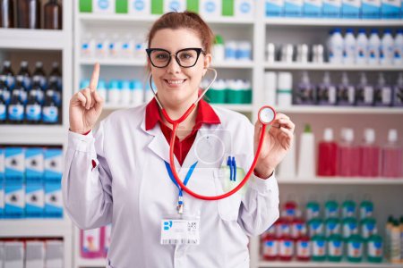 Photo for Young hispanic woman with red hair working at pharmacy drugstore using stethoscope smiling with an idea or question pointing finger with happy face, number one - Royalty Free Image