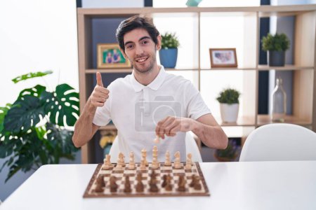 Téléchargez les photos : Jeune homme hispanique jouant aux échecs assis sur la table souriant heureux et positif, pouce levé faire excellent et signe d'approbation - en image libre de droit