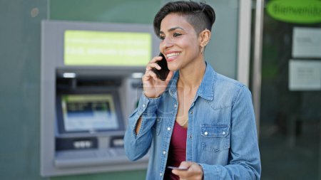 Téléchargez les photos : Jeune belle femme hispanique parlant sur smartphone tenant la carte de crédit au guichet de la banque - en image libre de droit
