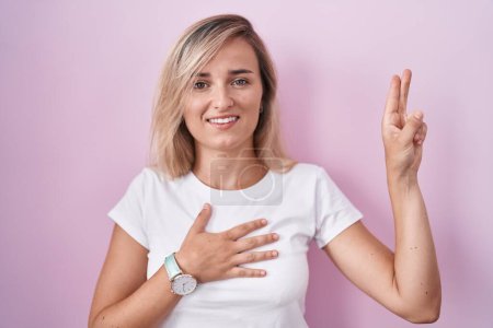 Foto de Mujer rubia joven de pie sobre fondo rosa sonriendo jurando con la mano en el pecho y los dedos hacia arriba, haciendo una promesa de lealtad juramento - Imagen libre de derechos