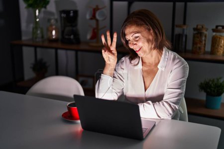 Photo for Middle age hispanic woman using laptop at home at night showing and pointing up with fingers number three while smiling confident and happy. - Royalty Free Image