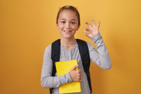 Photo for Little caucasian boy wearing student backpack and holding book doing ok sign with fingers, smiling friendly gesturing excellent symbol - Royalty Free Image