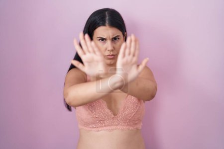 Foto de Mujer hispana joven con expresión de rechazo de sostén rosa cruzando brazos y palmas haciendo signo negativo, cara enojada - Imagen libre de derechos