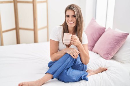 Foto de Mujer joven bebiendo taza de café sentado en la cama en el dormitorio - Imagen libre de derechos