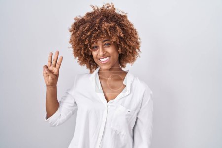 Foto de Mujer hispana joven con el pelo rizado de pie sobre fondo blanco mostrando y señalando hacia arriba con los dedos número tres mientras sonríe confiado y feliz. - Imagen libre de derechos