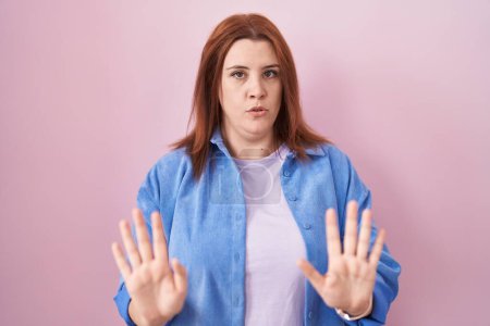 Foto de Mujer hispana joven con el pelo rojo de pie sobre fondo rosa alejando las palmas de las manos mostrando rechazo y negación con miedo y expresión repugnante. parada y prohibido. - Imagen libre de derechos