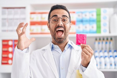 Foto de Hombre hispano con barba trabajando en farmacia sosteniendo condón celebrando la victoria con sonrisa feliz y expresión ganadora con las manos levantadas - Imagen libre de derechos