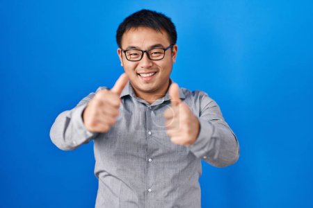 Téléchargez les photos : Jeune homme chinois debout sur fond bleu approuvant faire un geste positif avec la main, pouces levés souriant et heureux pour le succès. geste gagnant. - en image libre de droit