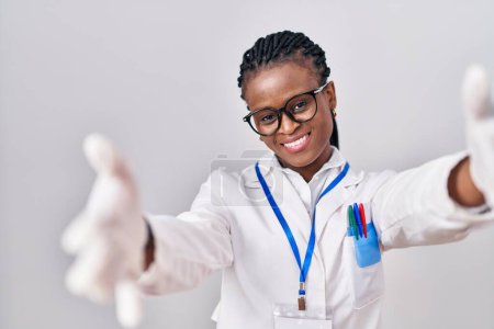 Photo for African woman with braids wearing scientist uniform looking at the camera smiling with open arms for hug. cheerful expression embracing happiness. - Royalty Free Image
