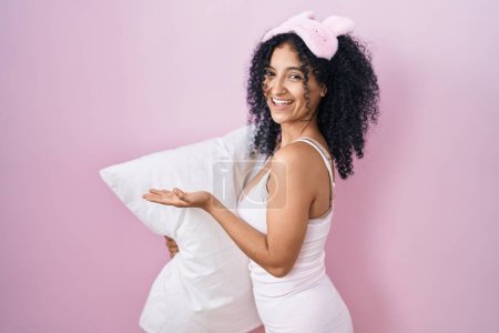 Foto de Mujer hispana con el pelo rizado con máscara de sueño y pijama sosteniendo almohada señalando a un lado con las manos abiertas mostrando espacio de copia, presentando anuncio sonriendo emocionada feliz - Imagen libre de derechos