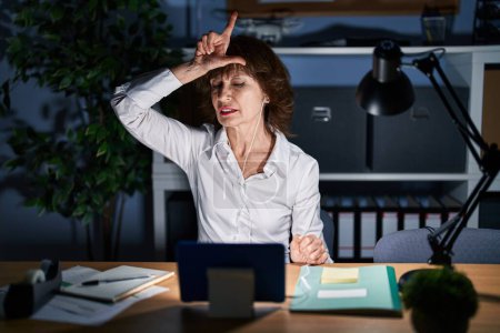 Photo for Middle age woman working at the office at night making fun of people with fingers on forehead doing loser gesture mocking and insulting. - Royalty Free Image