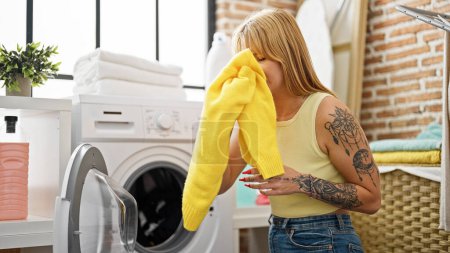 Foto de Mujer rubia joven lavando ropa oliendo ropa sucia en la lavandería - Imagen libre de derechos