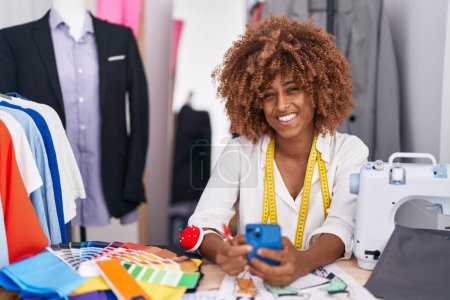 Téléchargez les photos : Femme afro-américaine tailleur souriant confiant en utilisant smartphone à l'atelier - en image libre de droit